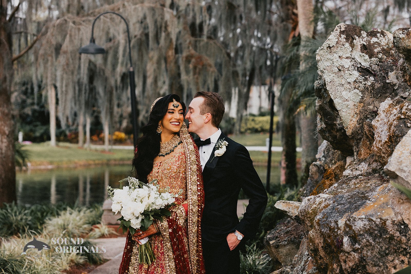 couple posing for photo in lush, natural environment. moss hangs from the trees and a rock formation sits behind them. the couple faces each other, the bride holding a bouquet of white flowers.